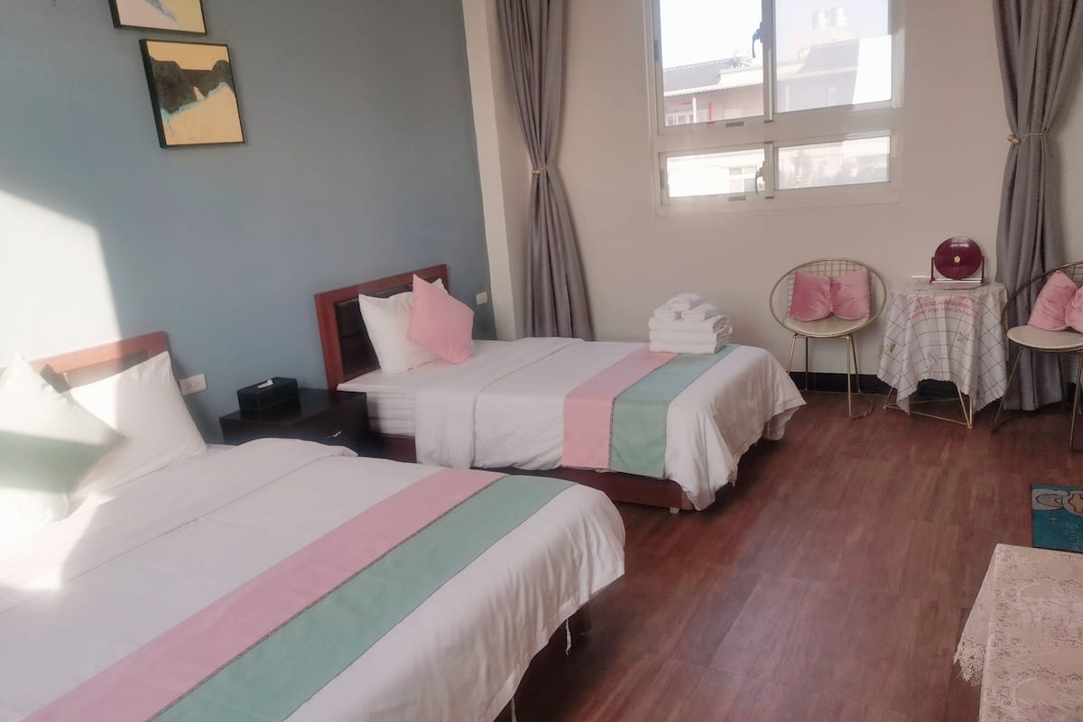 A hotel room with two beds, a small table with a red object, and a chair with pink cushions.