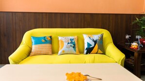 Un sofá amarillo brillante con tres cojines estampados, frente a una pared de madera y una mesa blanca con un jarrón de flores.