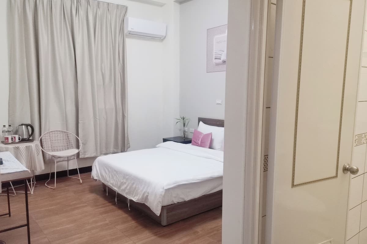 A bedroom with a bed, a desk with a chair, a nightstand, and a window with curtains.