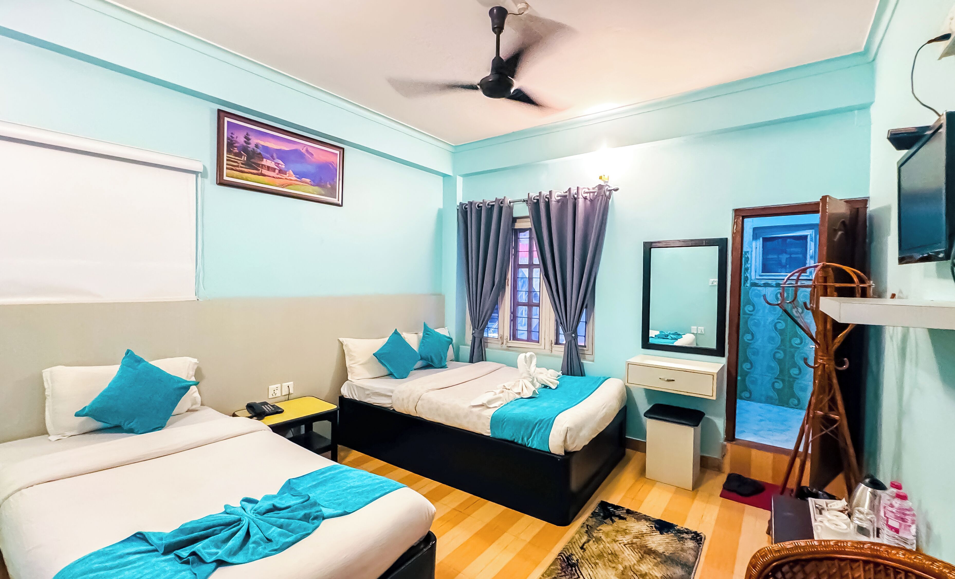 A hotel room with two beds, a desk, a chair, a mirror, a ceiling fan, and a window with curtains.