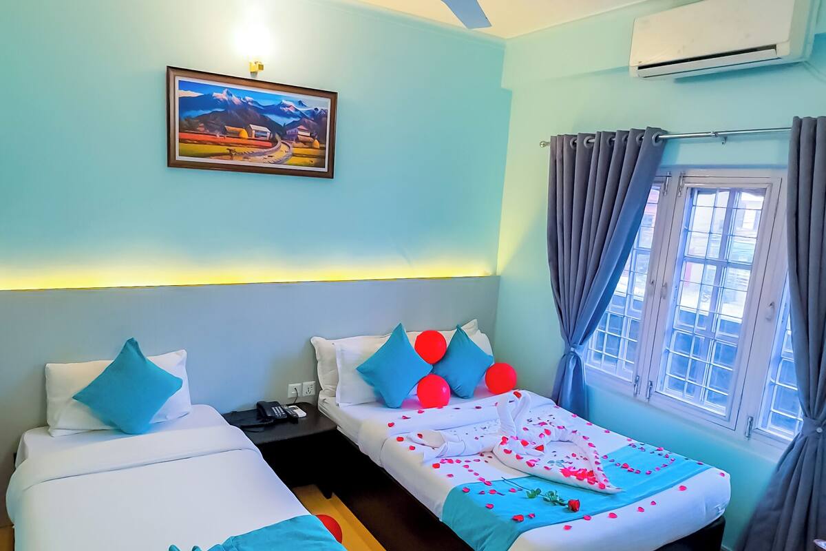 Habitación de hotel con dos camas, ventilador de techo, equipo de aire acondicionado, ventana con cortinas y un cuadro en la pared.