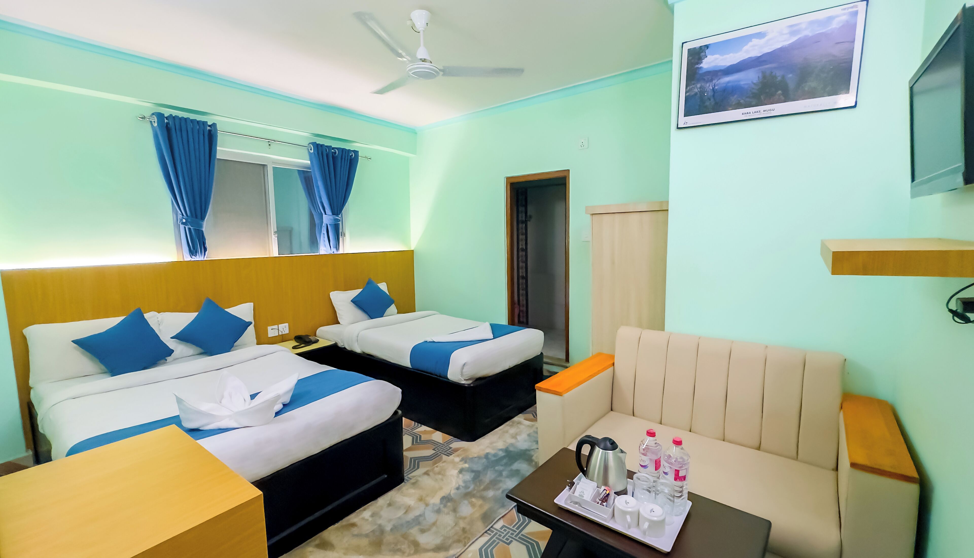 Habitación de hotel con dos camas, un sofá, una mesa de centro con artículos de cortesía, un ventilador de techo y un cuadro en la pared.