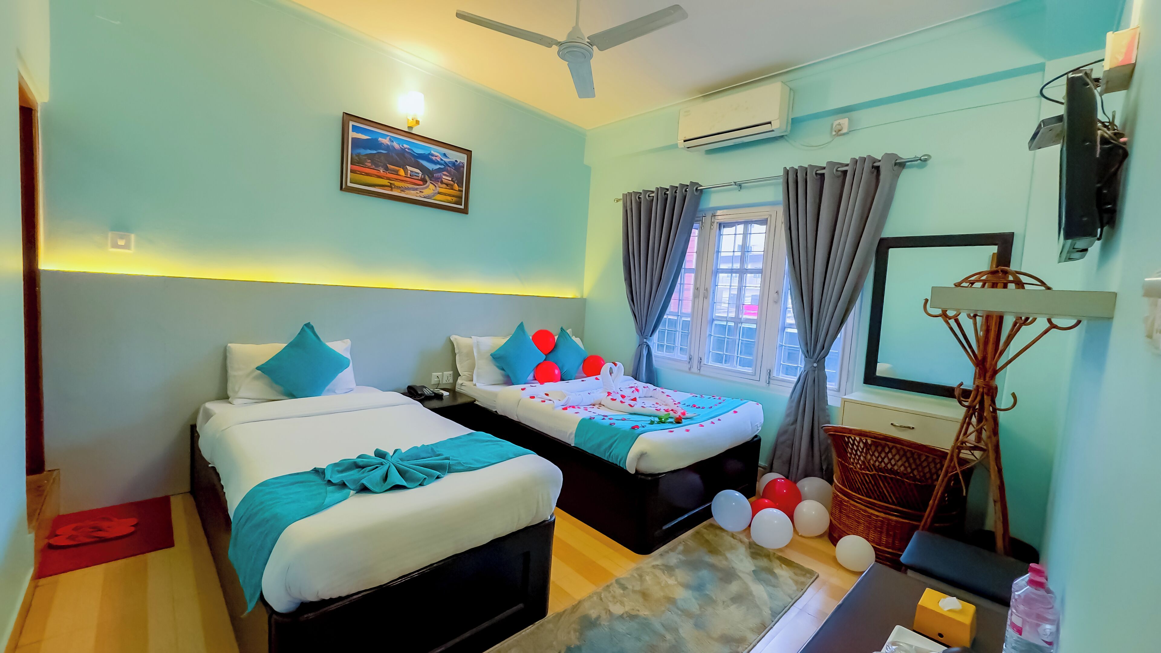 A hotel room with two beds, a ceiling fan, an air conditioning unit, a window with curtains, a wall-mounted TV, and a decorative basket.