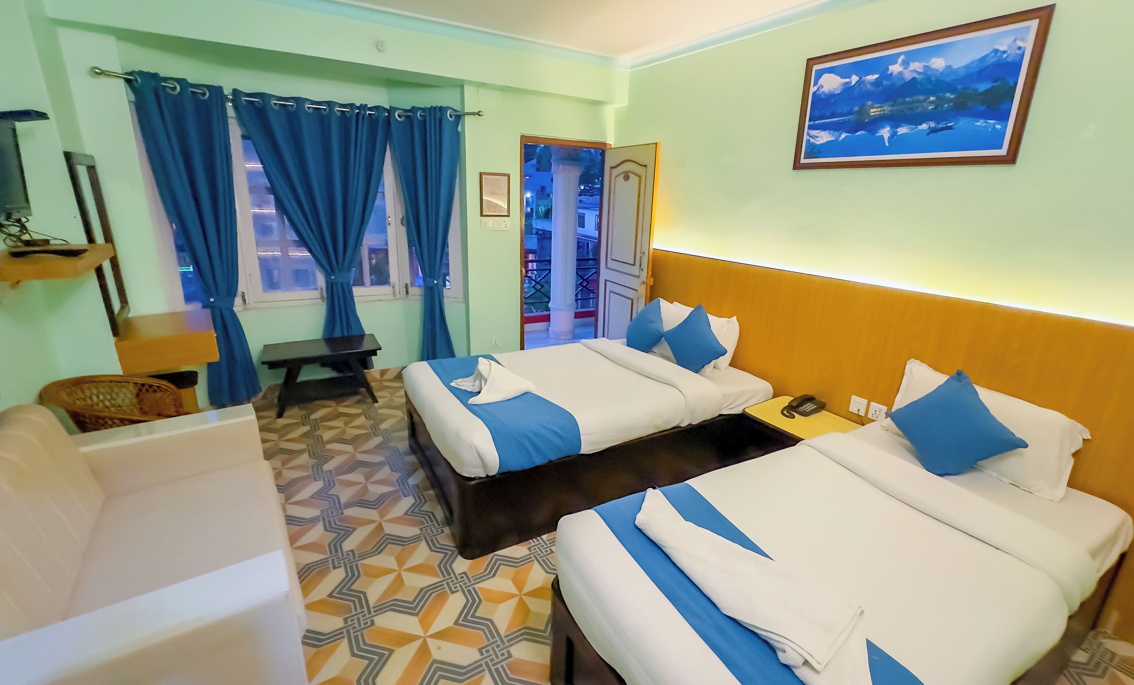 A hotel room with two beds, a desk, a chair, a window with curtains, a painting on the wall, and a patterned floor.