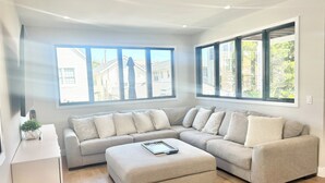 A modern living room with a sectional sofa, a small ottoman, and large windows offering a view of the neighborhood.