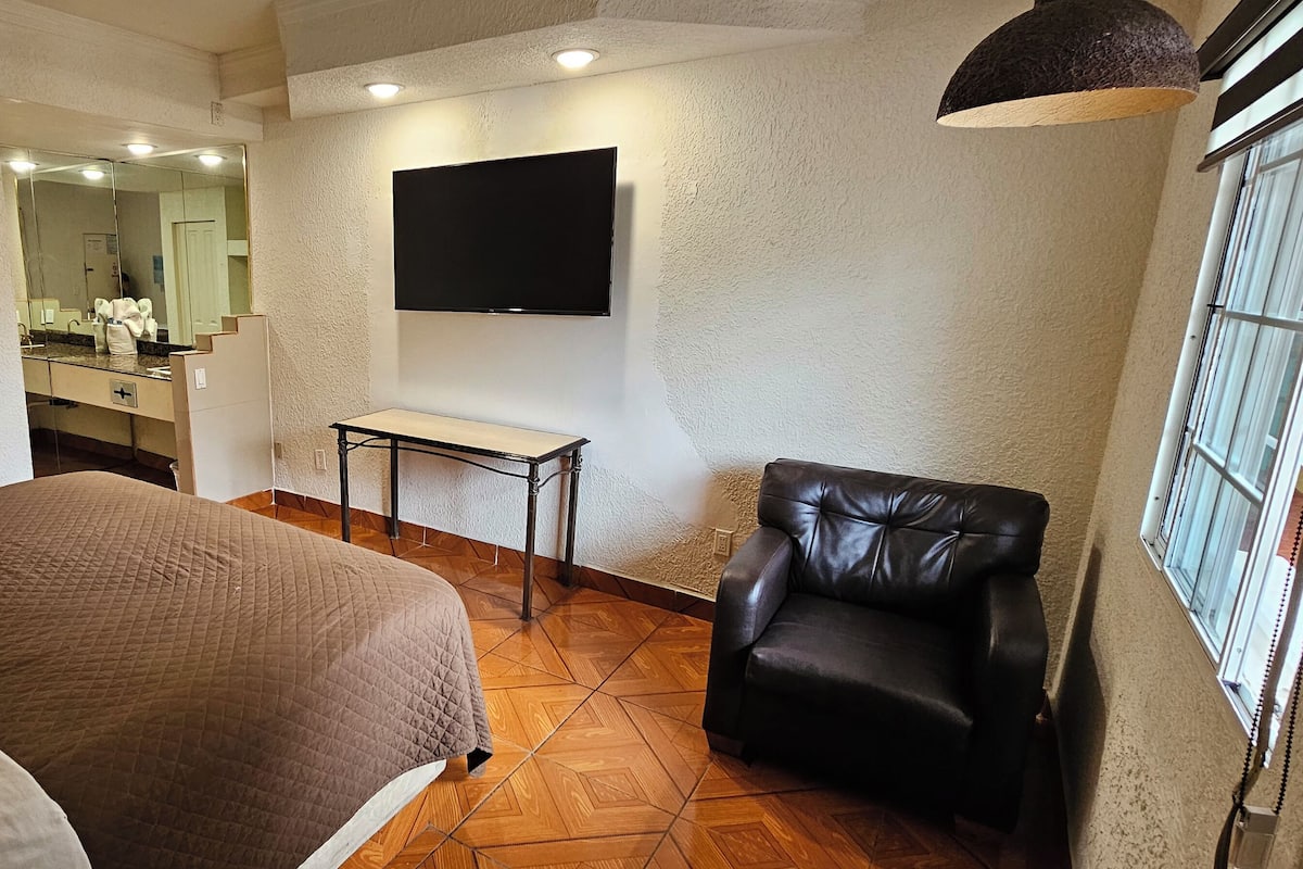 Un dormitorio con cama, escritorio, silla, televisor y baño visible a través de una puerta abierta.