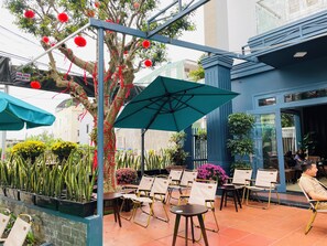 An outdoor seating area with chairs and tables, a tree with red decorations, and a blue umbrella. - Bossa Nova (Da Nang)