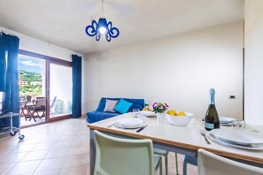 Uma área de refeições com mesa posta para dois, um sofá azul e uma varanda com vista.
