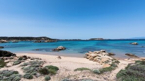 Ein Strand mit klarem türkisblauem Wasser, Felsformationen und spärlicher Vegetation.