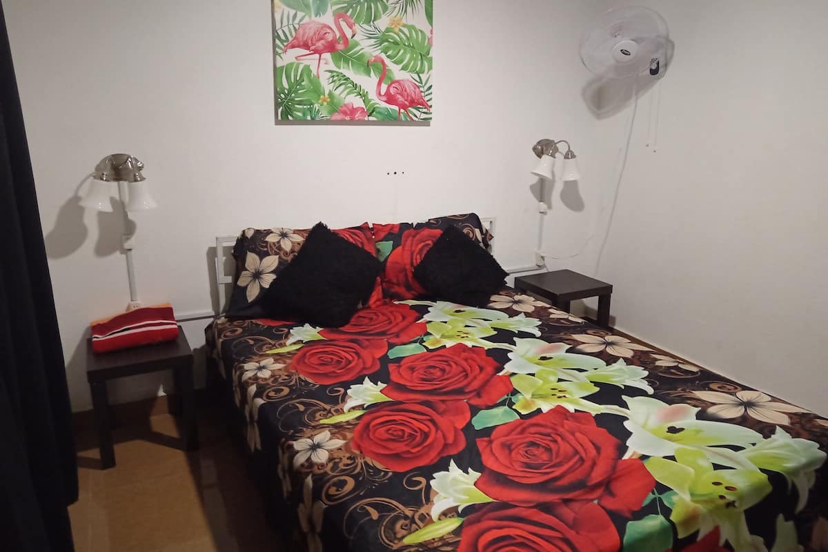 A bedroom with a bed, two bedside lamps, a nightstand, a wall-mounted fan, and a painting of flamingos and leaves.
