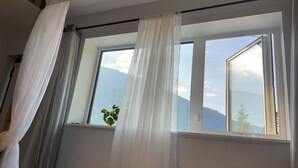 Ett rum med ett stort fönster, vita gardiner och utsikt över bergen.