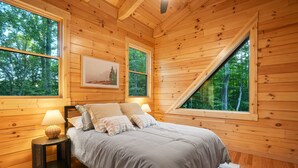 A bedroom with a bed, two bedside lamps, a framed picture on the wall, and large windows with a view of trees. - Rustic Ravine | Hocking Hills, Hot Tub, Game Room (South Bloomingville)