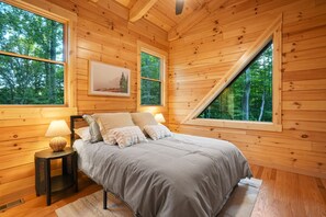 A wooden cabin bedroom with a bed, two windows, a nightstand, and a lamp. - Rustic Ravine | Hocking Hills, Hot Tub, Game Room (South Bloomingville)
