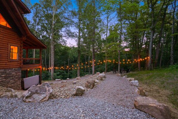 Une cabane en bois avec un chemin de gravier menant à une terrasse, entourée d’arbres et illuminée de guirlandes lumineuses.