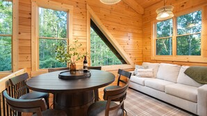 A wooden cabin interior with a dining table, chairs, a sofa, and large windows offering a view of the forest. - Rustic Ravine | Hocking Hills, Hot Tub, Game Room (South Bloomingville)