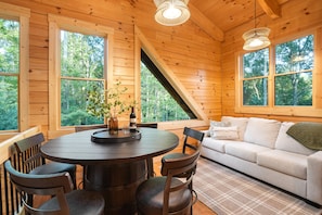 A wooden cabin interior with a round dining table, chairs, a sofa, and large windows offering a view of the forest.