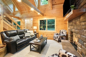 A cozy log cabin interior with a leather sofa, a coffee table, a fireplace, and a stone wall. - Rustic Ravine | Hocking Hills, Hot Tub, Game Room (South Bloomingville)