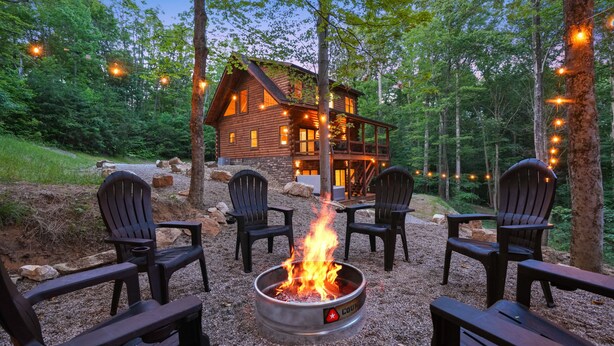 A cabin with a fire pit surrounded by Adirondack chairs, set in a forested area. - Rustic Ravine | Hocking Hills, Hot Tub, Game Room (South Bloomingville)