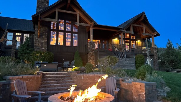 Una casa grande de estilo rústico con una chimenea destacada, asientos exteriores e ingreso bien iluminado.