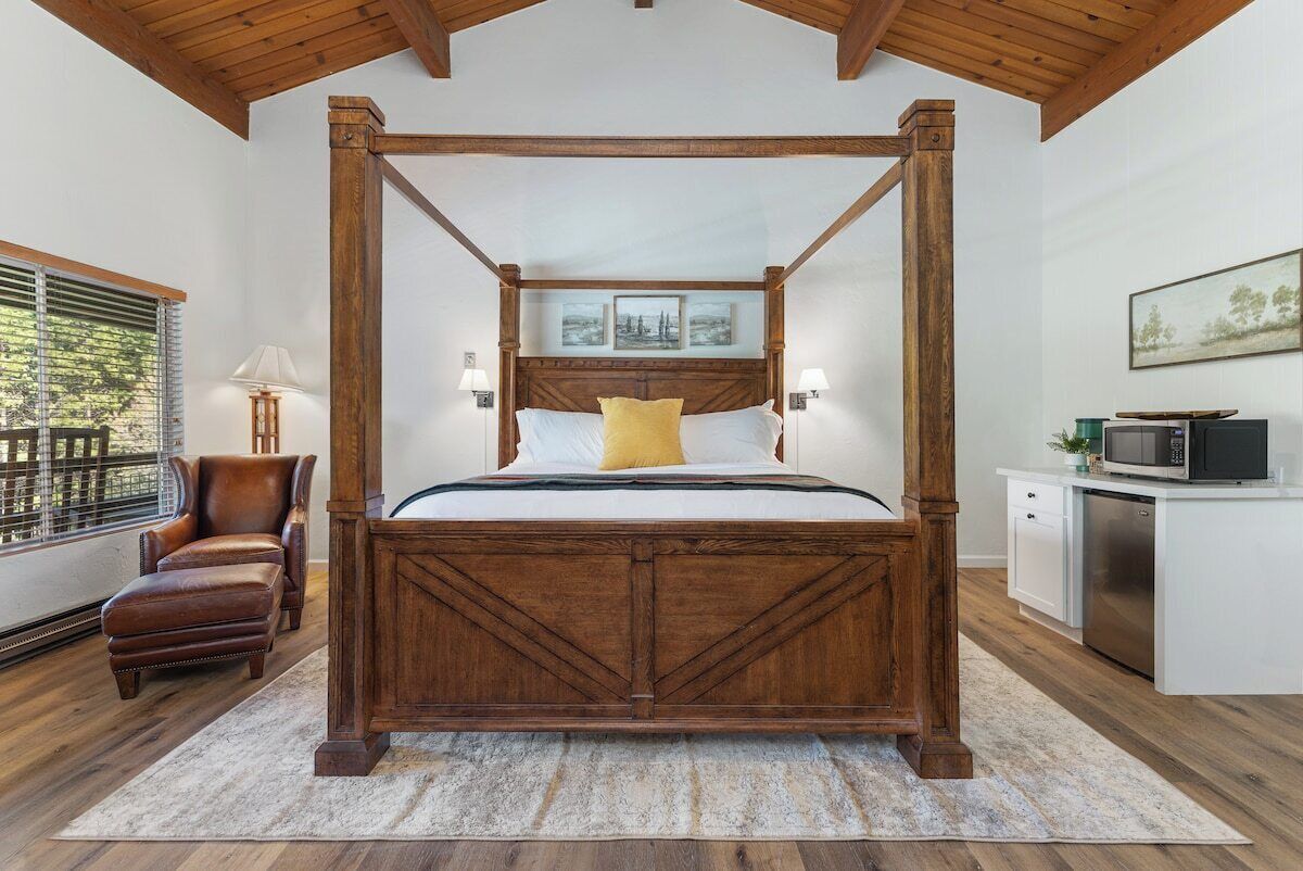 A four-poster bed with a wooden headboard, a brown leather chair, a small desk with a microwave, and a window with blinds.