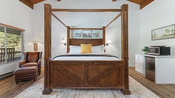 A four-poster bed with a wooden headboard, a brown leather chair, a small desk with a microwave, and a window with blinds.
