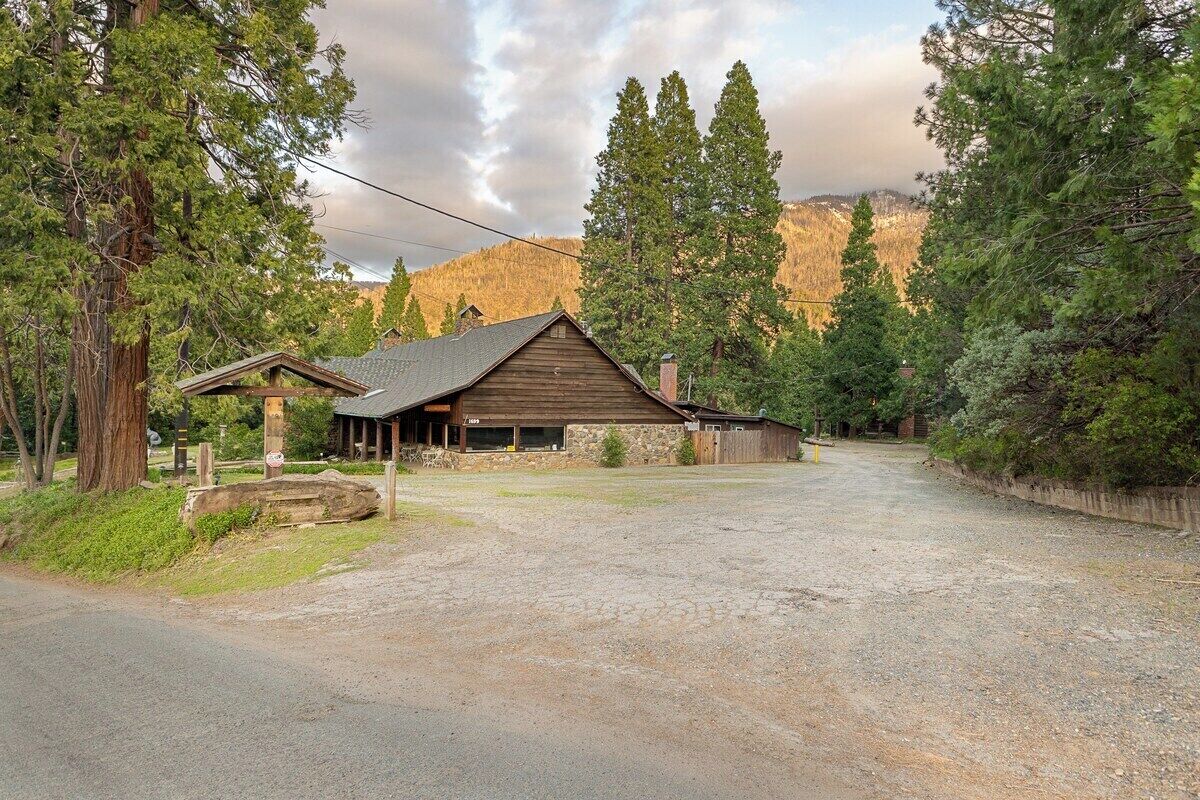A rustic cabin with a stone foundation, a wooden roof, and a gravel driveway leading to a wooden gate.