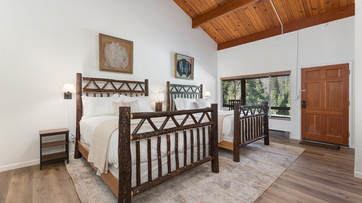 Quarto com duas camas, cabeceiras de madeira, uma pequena mesa de cabeceira, mesa de apoio e uma janela grande com persianas, além de uma porta de madeira.
