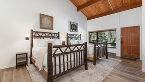 A bedroom with two beds, wooden headboards, a small bedside table, a nightstand, a large window with blinds, and a wooden door. - Camp Nelson Lodge Room 9 (Springville)