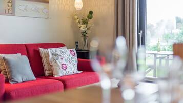 Ein rotes Sofa mit floralen Kissen, eine Vase mit Blumen und eine Pendelleuchte in einem Raum mit Blick auf Grünfläche.