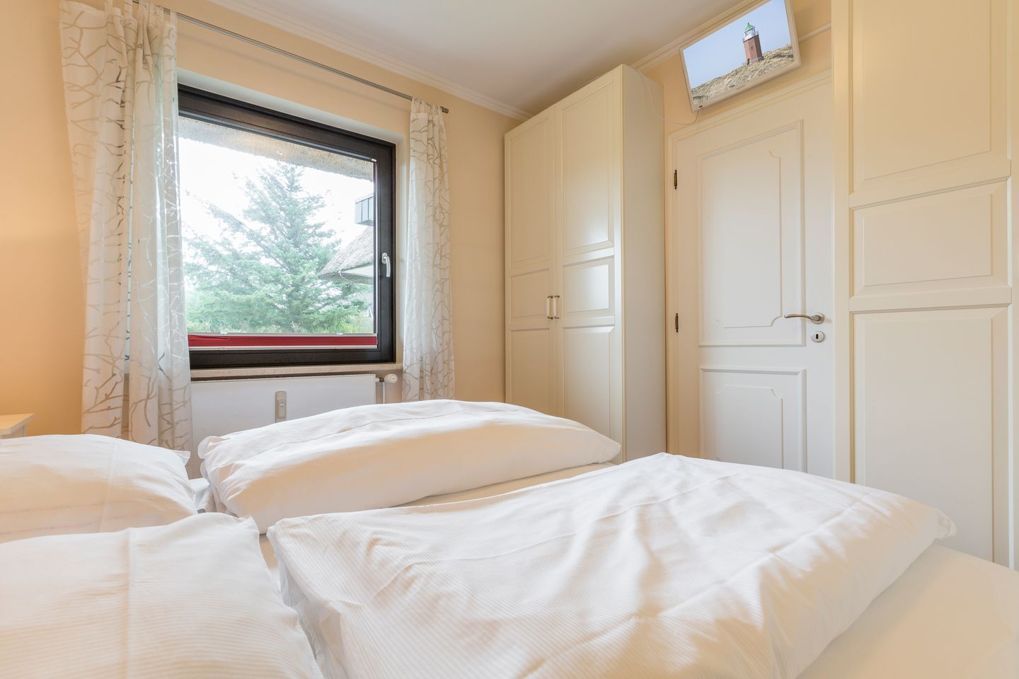 Ein Schlafzimmer mit zwei Betten, einem Fenster mit Vorhängen, einem Heizkörper und einem an der Wand befestigten Fernseher.