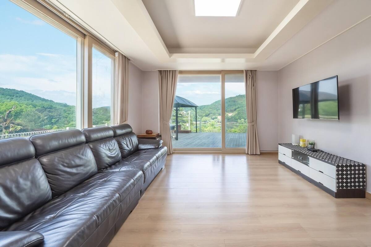 A spacious living room with a leather sofa, a TV mounted on the wall, and a large sliding glass door leading to a balcony with a scenic view.