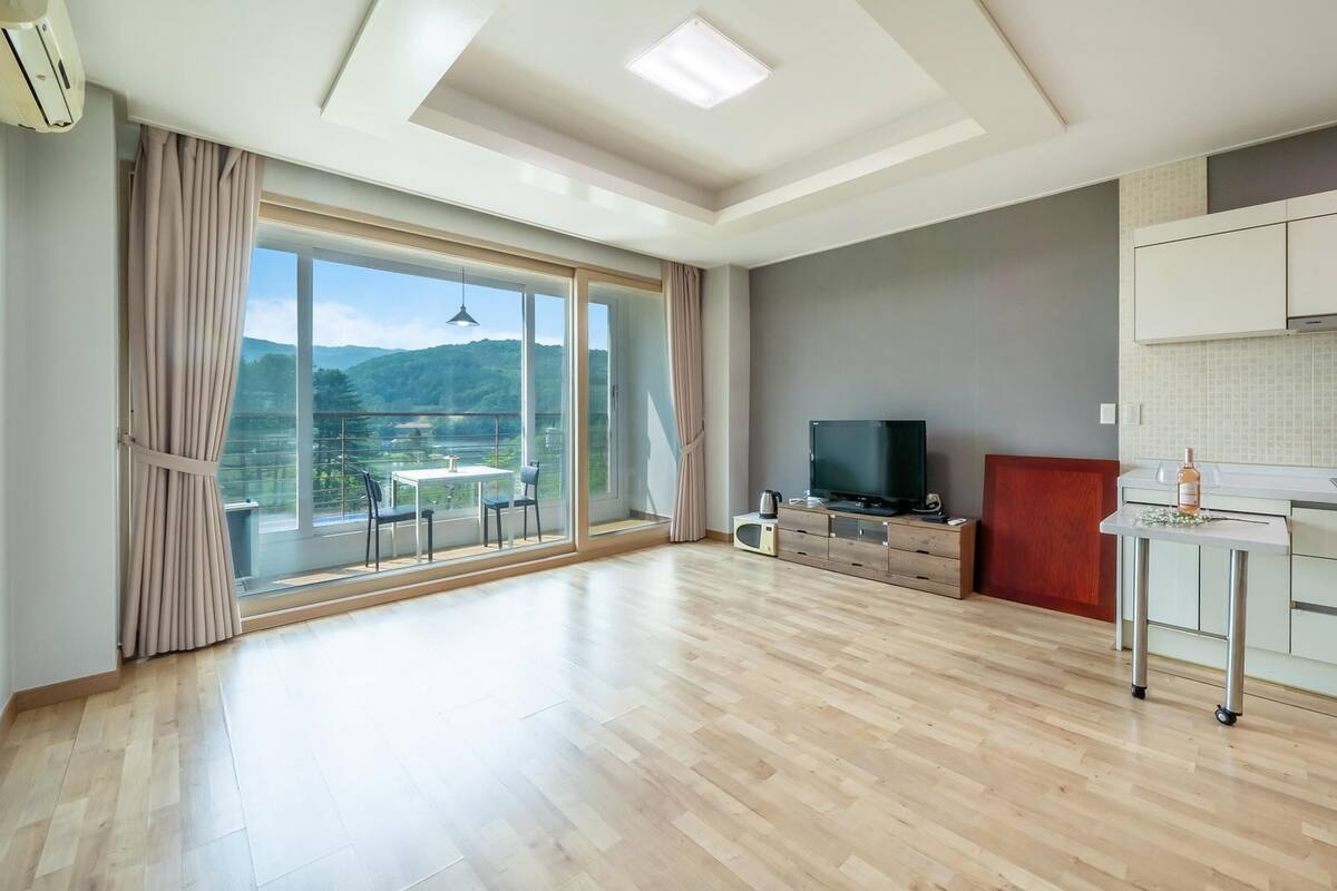 A spacious living area with a large sliding glass door leading to a balcony, a flat-screen TV, a small kitchen area, and a dining table with chairs.