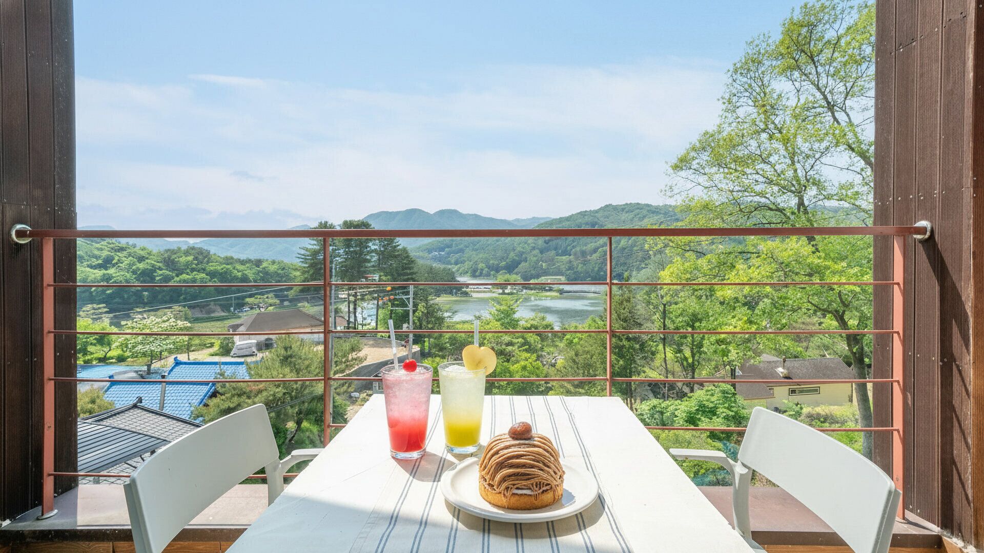A balcony with a table set for two, overlooking a scenic landscape with a river and hills.