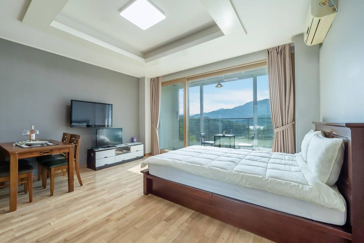 A modern bedroom with a large bed, a TV, a dining table, and a balcony with a mountain view.