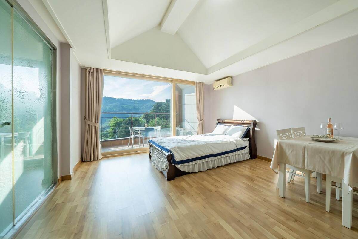 A bedroom with a bed, a dining table with chairs, a balcony with a table and chairs, and a view of the outdoors.