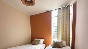 Ein Hotelzimmer mit zwei Einzelbetten, einer gewobenen Deckenleuchte, einem Fenster mit Vorhängen und einem Holzboden mit Fischgrätmuster.