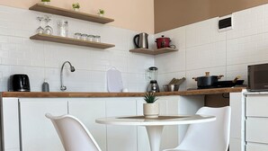 Eine kleine Küche mit weißen Schränken, einem runden Esstisch mit zwei weißen Stühlen und einer Arbeitsplatte mit verschiedenen Küchengeräten und Utensilien.