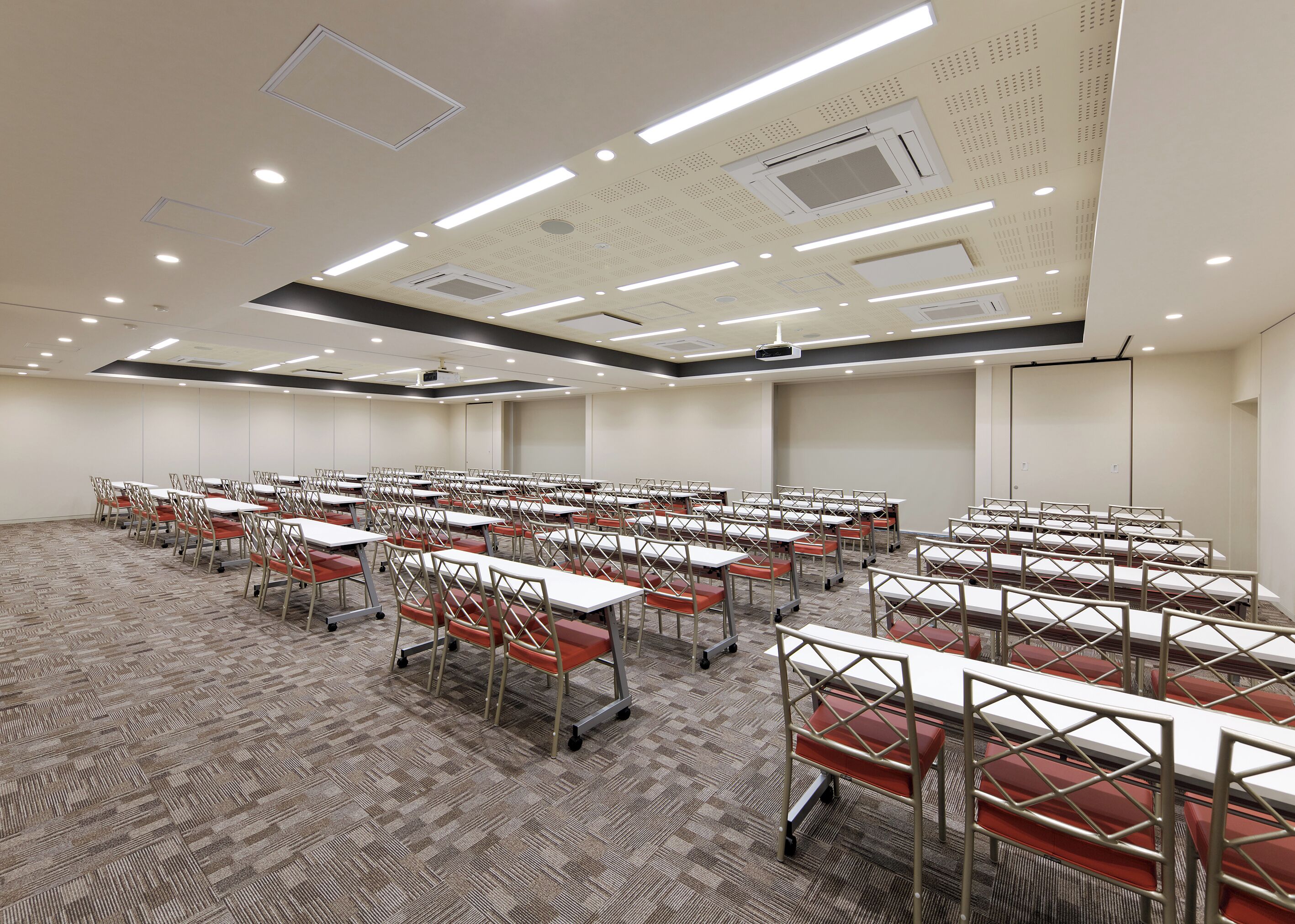 A large conference room with rows of tables and chairs, equipped with ceiling projectors and ventilation.