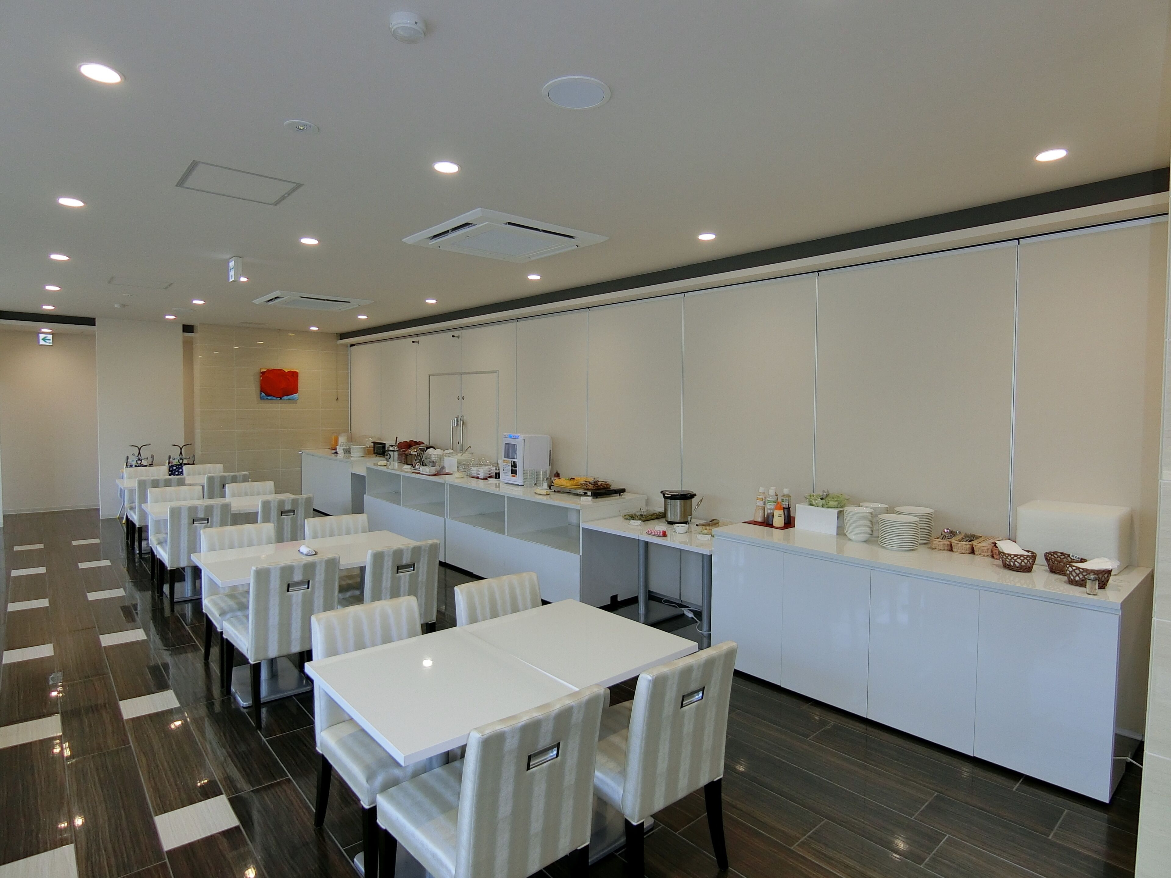 A modern hotel room with a dining area featuring white tables and chairs, a kitchen counter with various appliances, and a tiled floor.