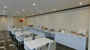 A modern hotel room with a dining area featuring white tables and chairs, a kitchen counter with various appliances, and a tiled floor.
