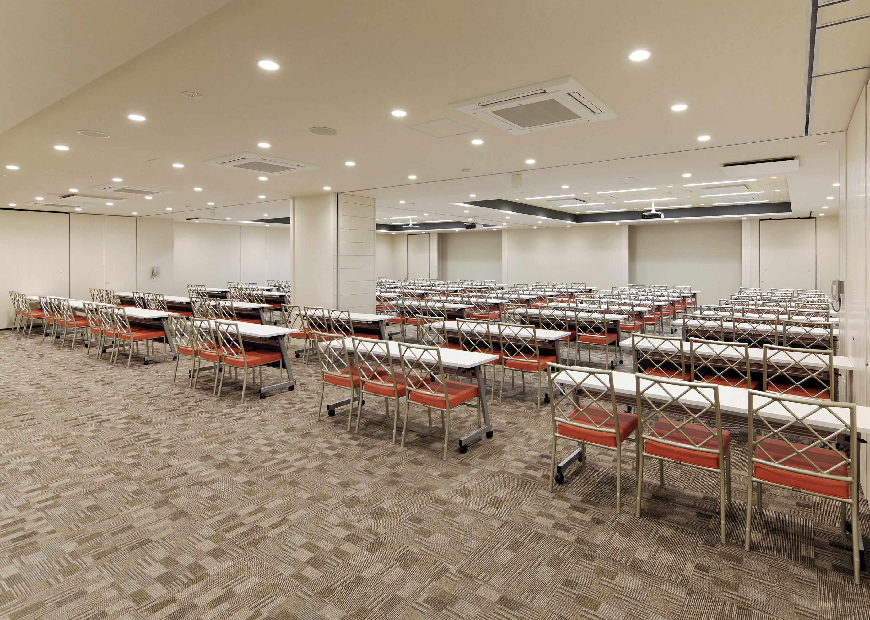 A large conference room with rows of tables and chairs, carpeted flooring, and recessed ceiling lighting.