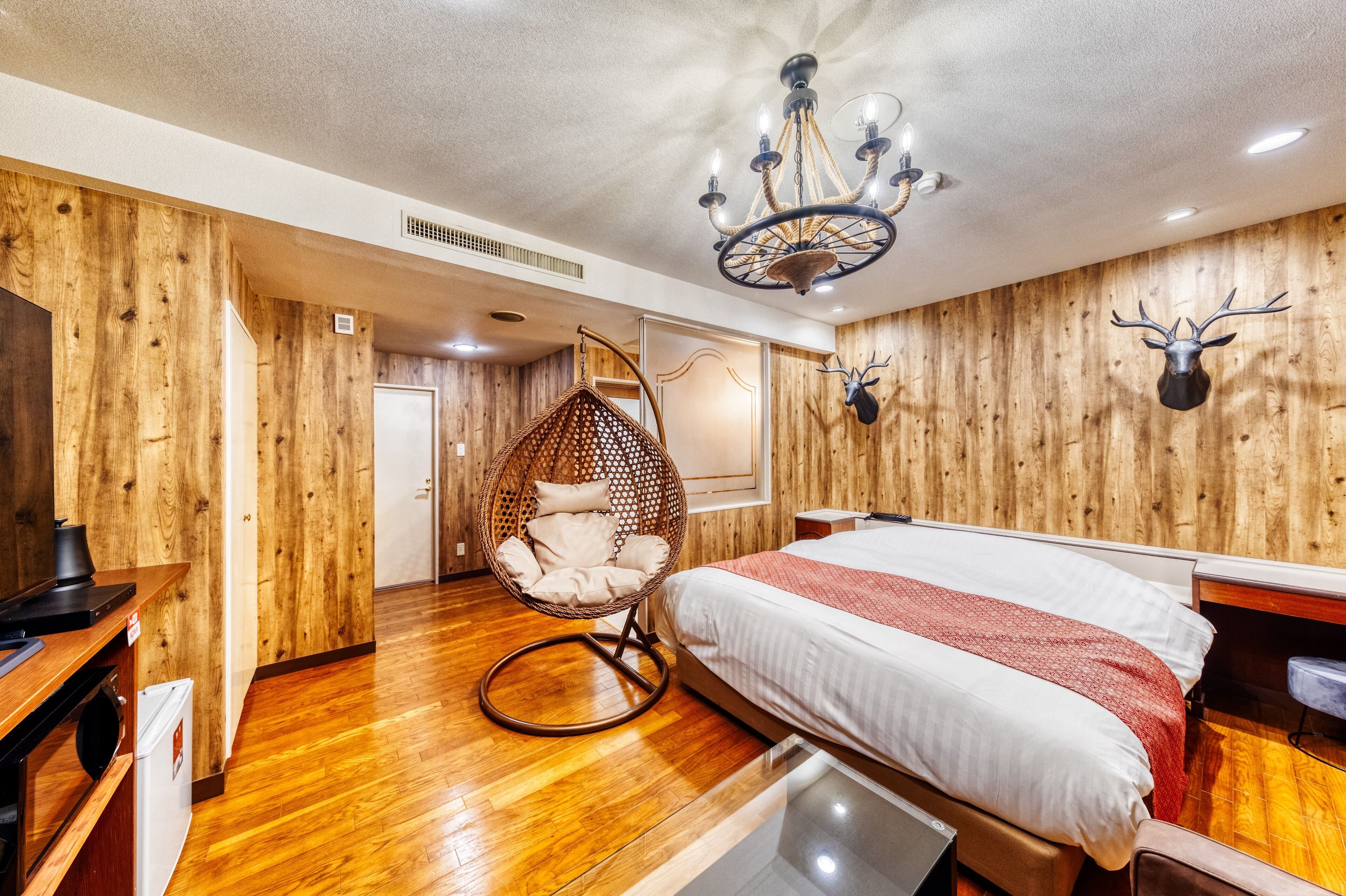Una habitación con piso de madera, una cama con manta roja, una silla colgante y un candelabro.