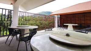 Un balcón con dos mesas redondas y sillas, baranda metálica y vistas de vegetación exuberante y montañas.