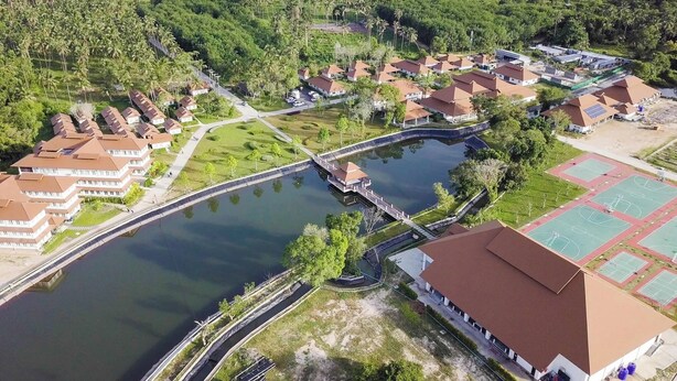 Vista aérea de un resort con varias cabañas tipo bungalow, un elemento acuático central y una zona de canchas deportivas.