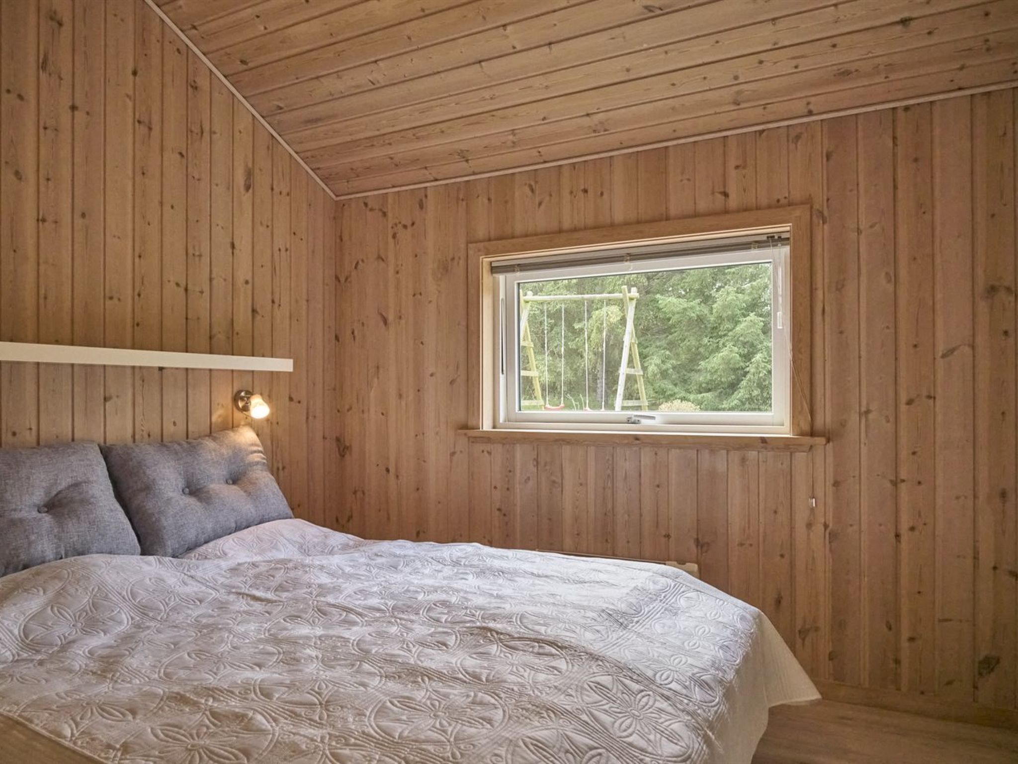 A wooden-paneled bedroom with a bed, a window with a view of trees, and a wall-mounted light fixture.