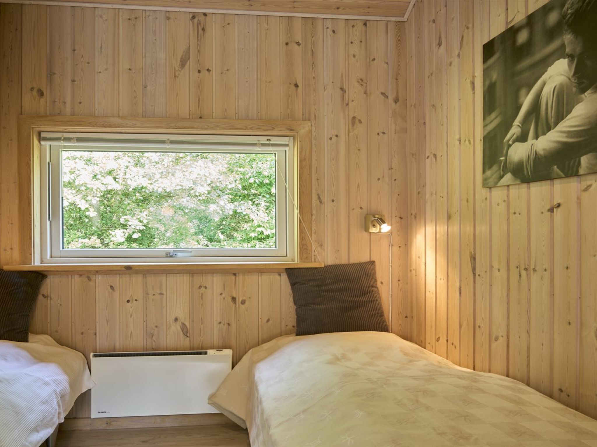 A wooden-paneled room with two beds, a window with a view of greenery, and a wall-mounted light fixture.