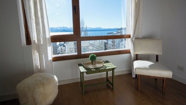 Ein Zimmer mit großem Fenster, einem kleinen Tisch mit Pflanze, einem weißen Stuhl und einem kuscheligen Hocker.