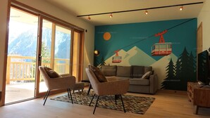 Ein Wohnzimmer mit großem Fenster, einer Couch, zwei Sesseln, einem gemusterten Teppich und einem Wandgemälde mit einer Berglandschaft und einer Seilbahn.