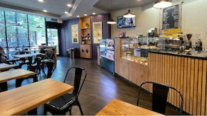 Interior de una cafetería con mesas de madera, sillas negras, una barra con una máquina de café y una pizarra de menú.