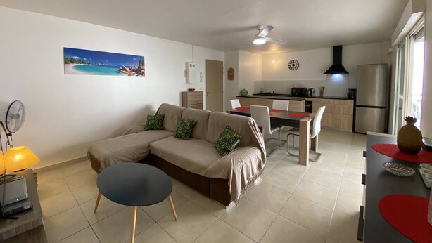 A modern living room with a beige sofa, a small round table, and a fan. A kitchen is visible in the background with a refrigerator, stove, and dining area. - Le Clos de l'Allamanda - Le Marin (LE MARIN)